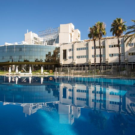 Silken Al-Andalus Palace Seville Exterior photo The photo features a modern hotel with a large glass facade reflecting in a calm blue swimming pool. The building is surrounded by palm trees, indicating a tropical or warm climate. The clear blue sky adds to the serene atmosphere, while lounge chair