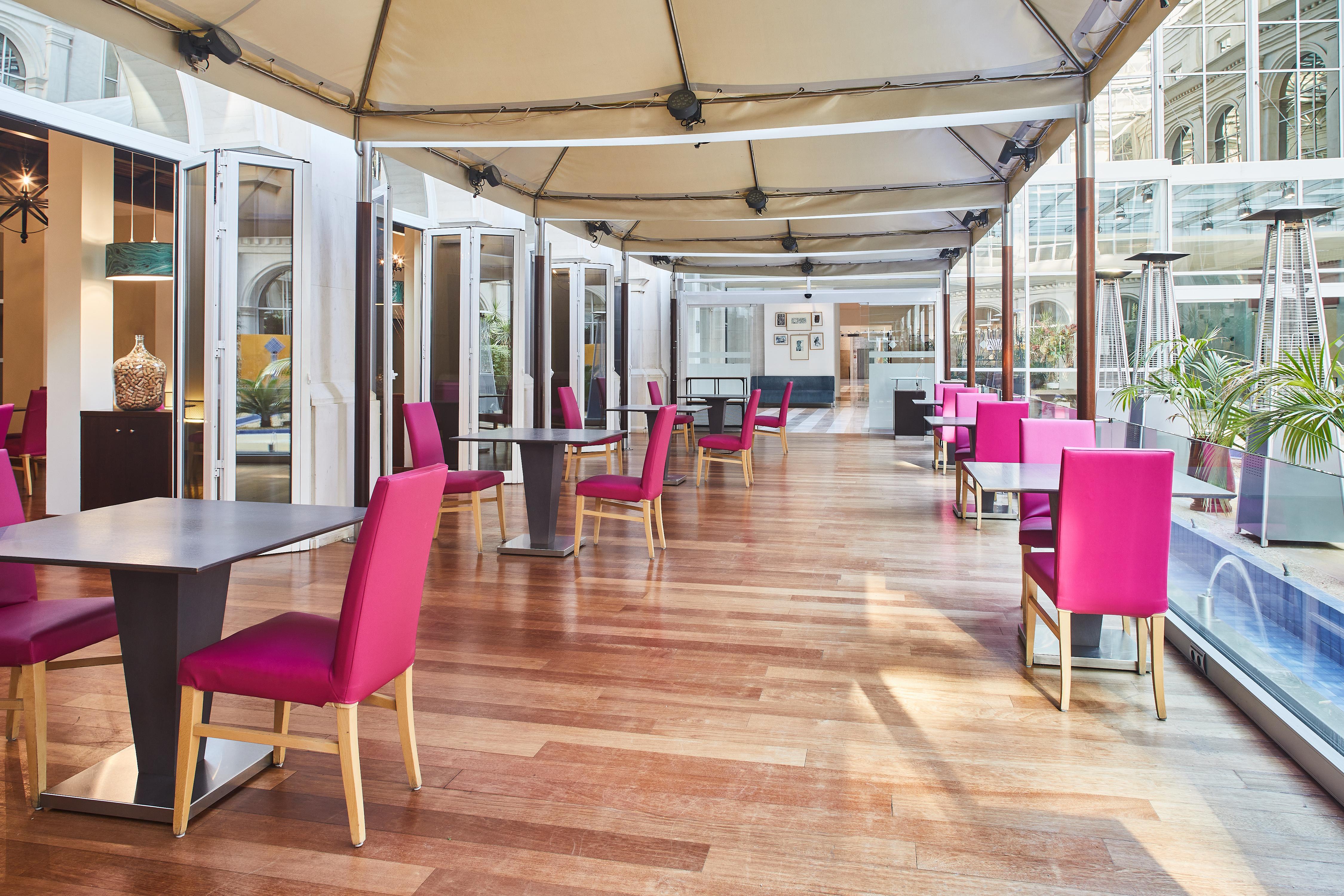 Silken Al-Andalus Palace Seville Exterior photo The photo shows a modern dining area with a sleek design. It features wooden flooring and tables set up in a row. Each table has a single pink chair positioned beside it. Large glass windows or doors line one side, creating an open and airy atmospher