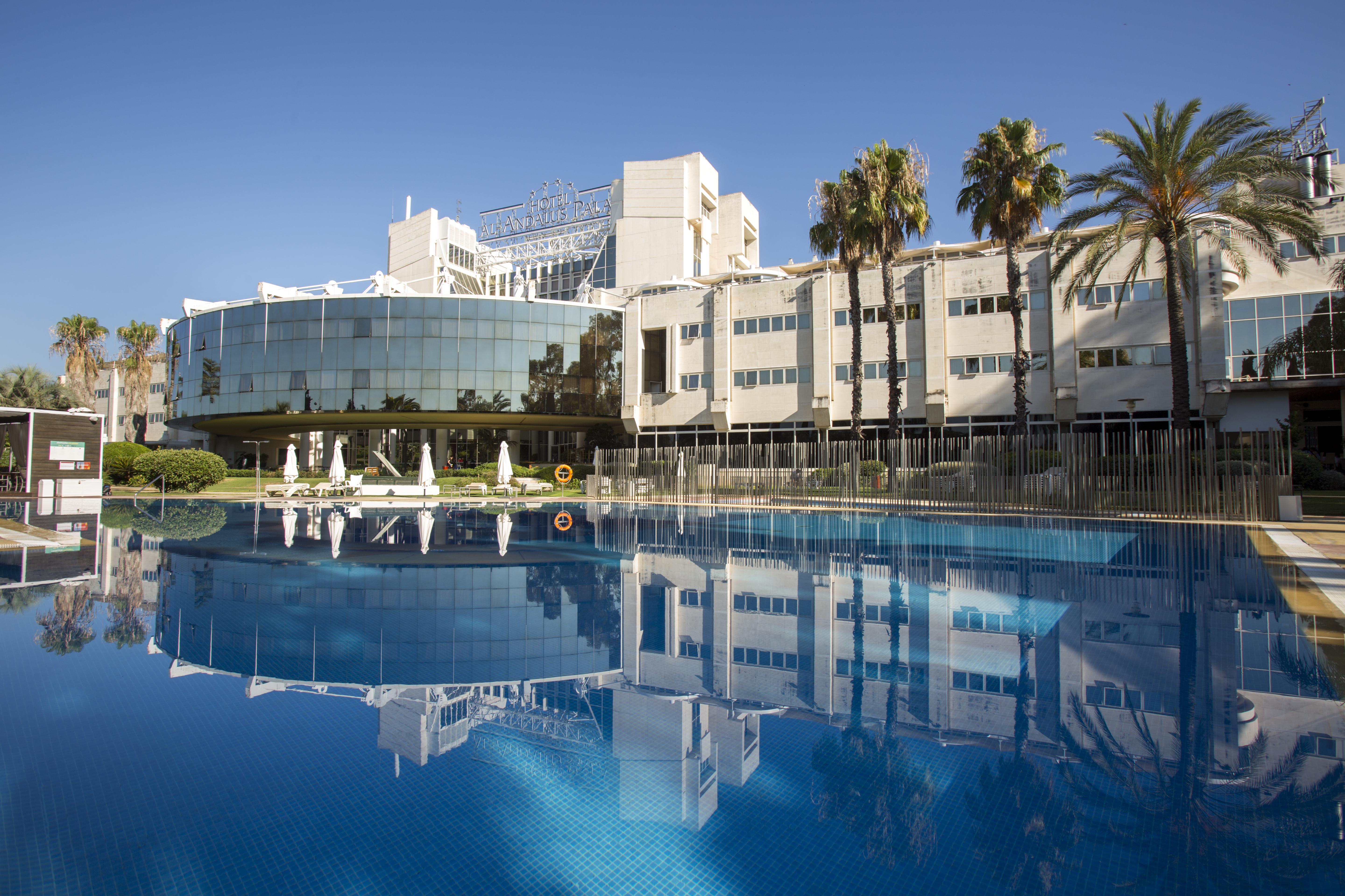 Silken Al-Andalus Palace Seville Exterior photo The photo features a modern hotel with a large glass facade reflecting in a calm blue swimming pool. The building is surrounded by palm trees, indicating a tropical or warm climate. The clear blue sky adds to the serene atmosphere, while lounge chair
