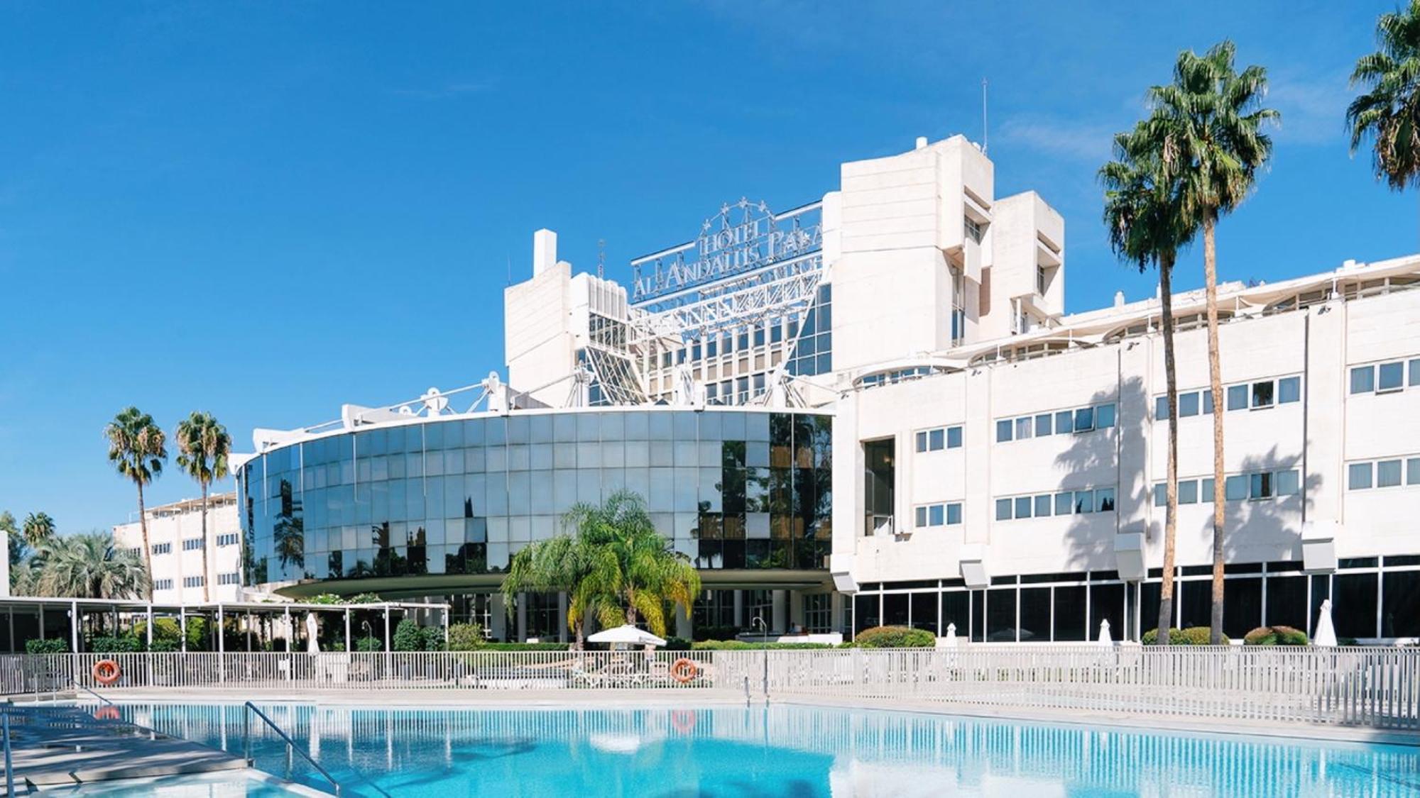 Silken Al-Andalus Palace Seville Exterior photo The photo shows a modern hotel building with a large glass facade, reflecting the surrounding area. There are palm trees in the foreground, and a swimming pool in the forefront of the image. The hotel has a stylish design, with a prominent sign on to